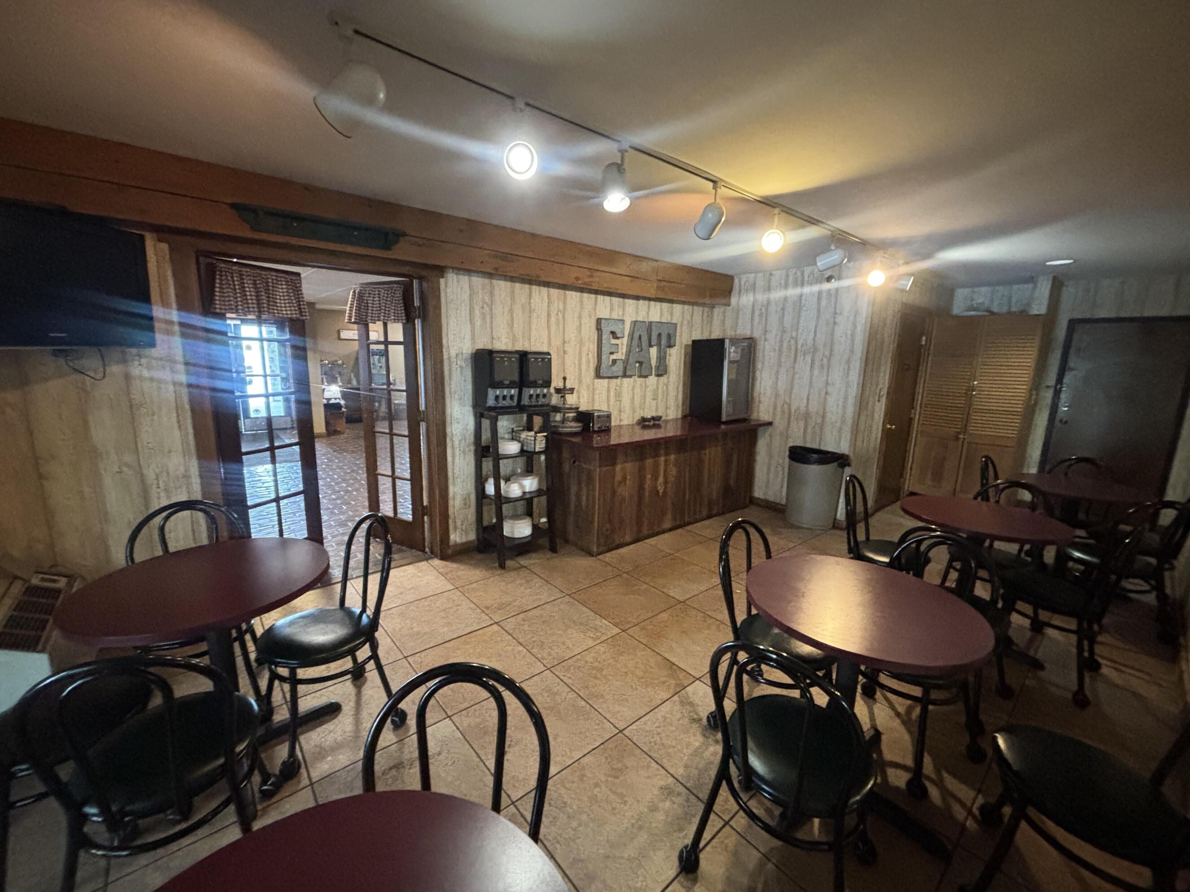 A restaurant interior with multiple tables and chairs, a counter with a beverage dispenser, and a view of the outside through a door.