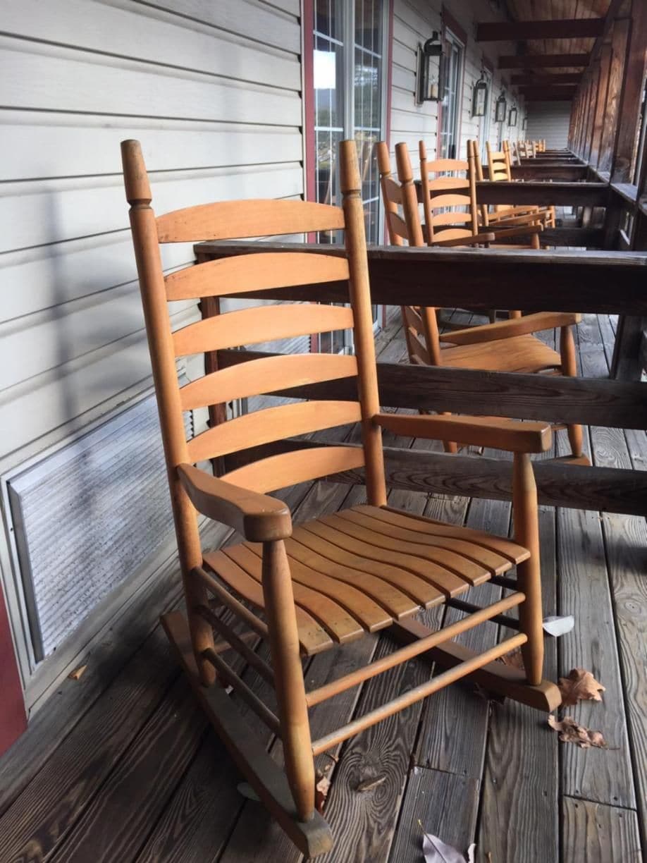 A row of wooden rocking chairs on a wooden deck with a building in the background.