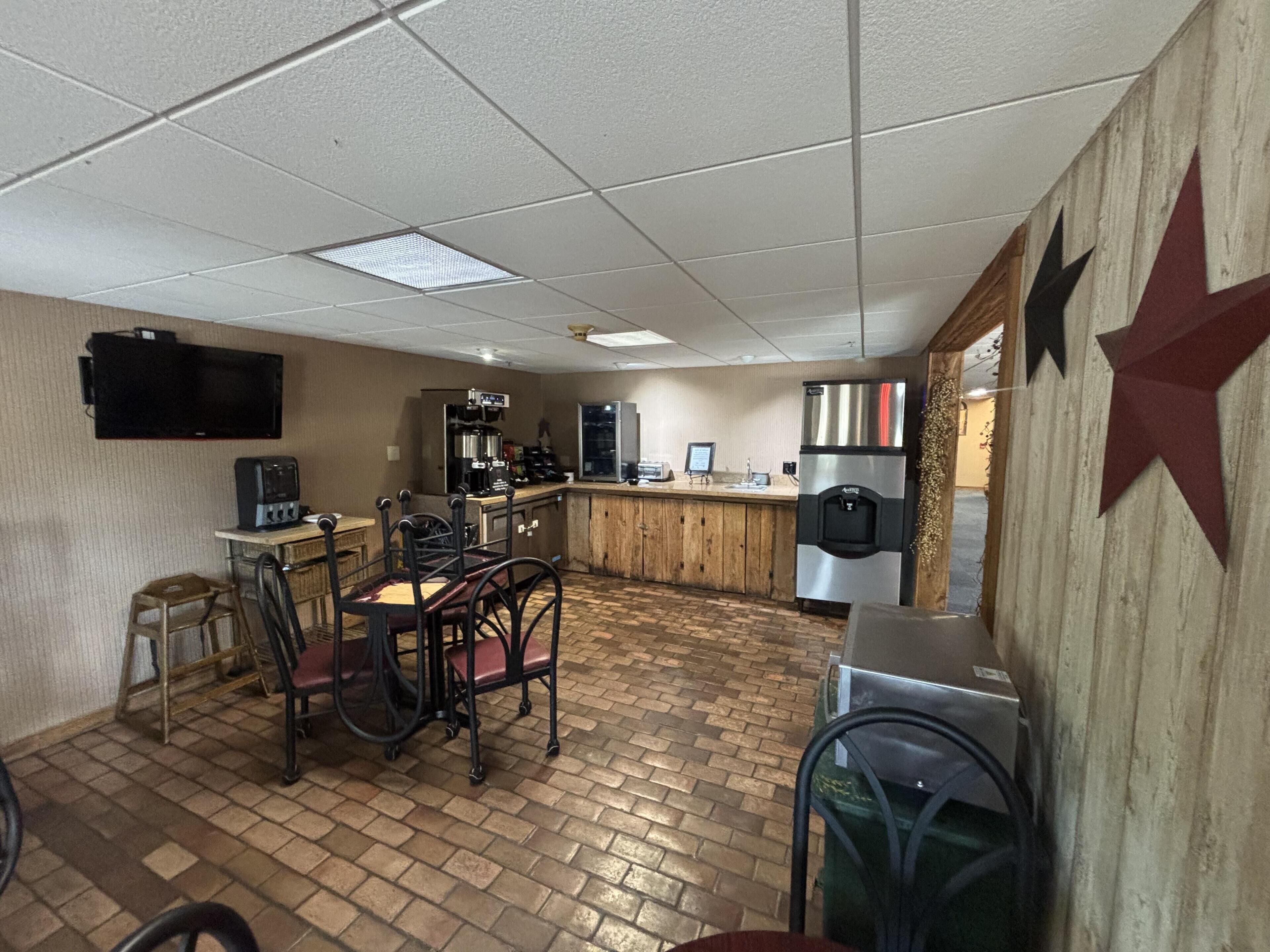 A kitchen area with a brick floor, a coffee machine, a refrigerator, and a television mounted on the wall.