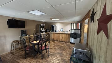 A kitchen area with a brick floor, a coffee machine, a refrigerator, and a television mounted on the wall.