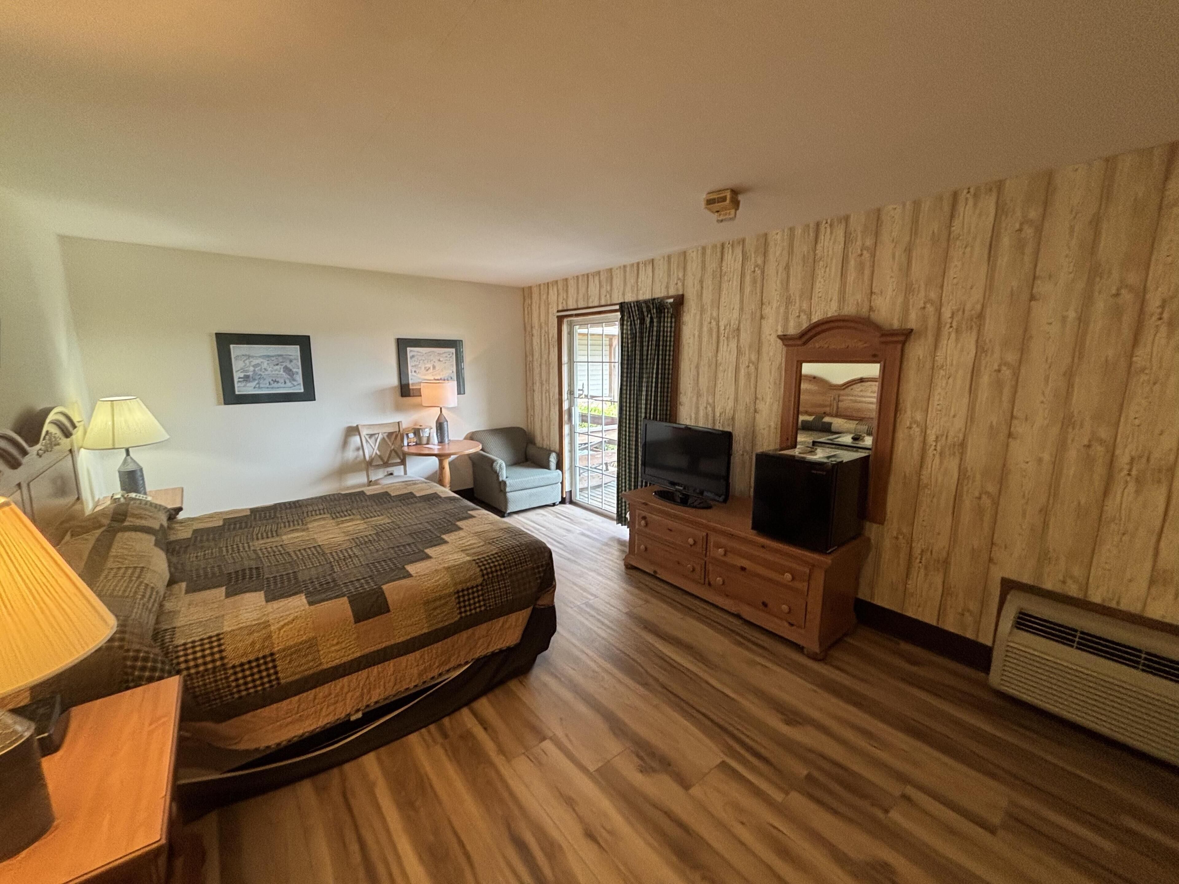 A hotel room with a bed, a dresser, a television, a chair, a table with a lamp, and a window with curtains.