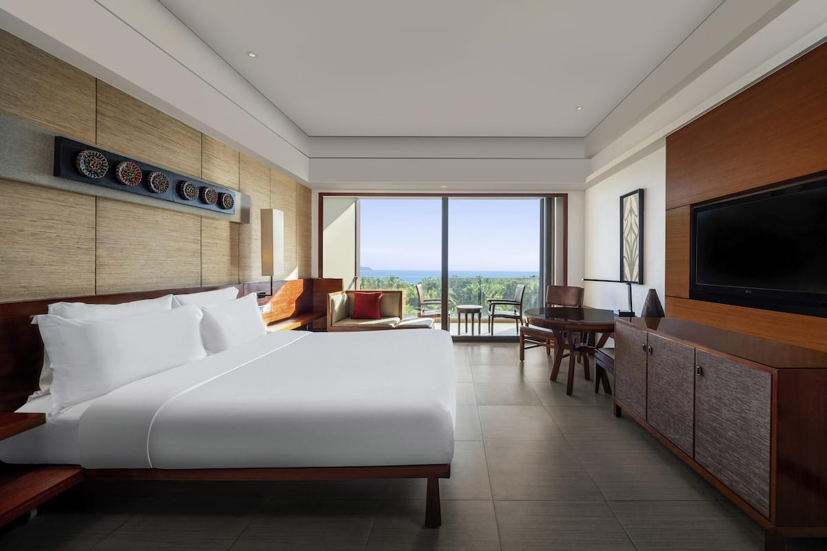 Quarto de hotel moderno com uma cama grande, televisão, área de refeições e varanda com vista.