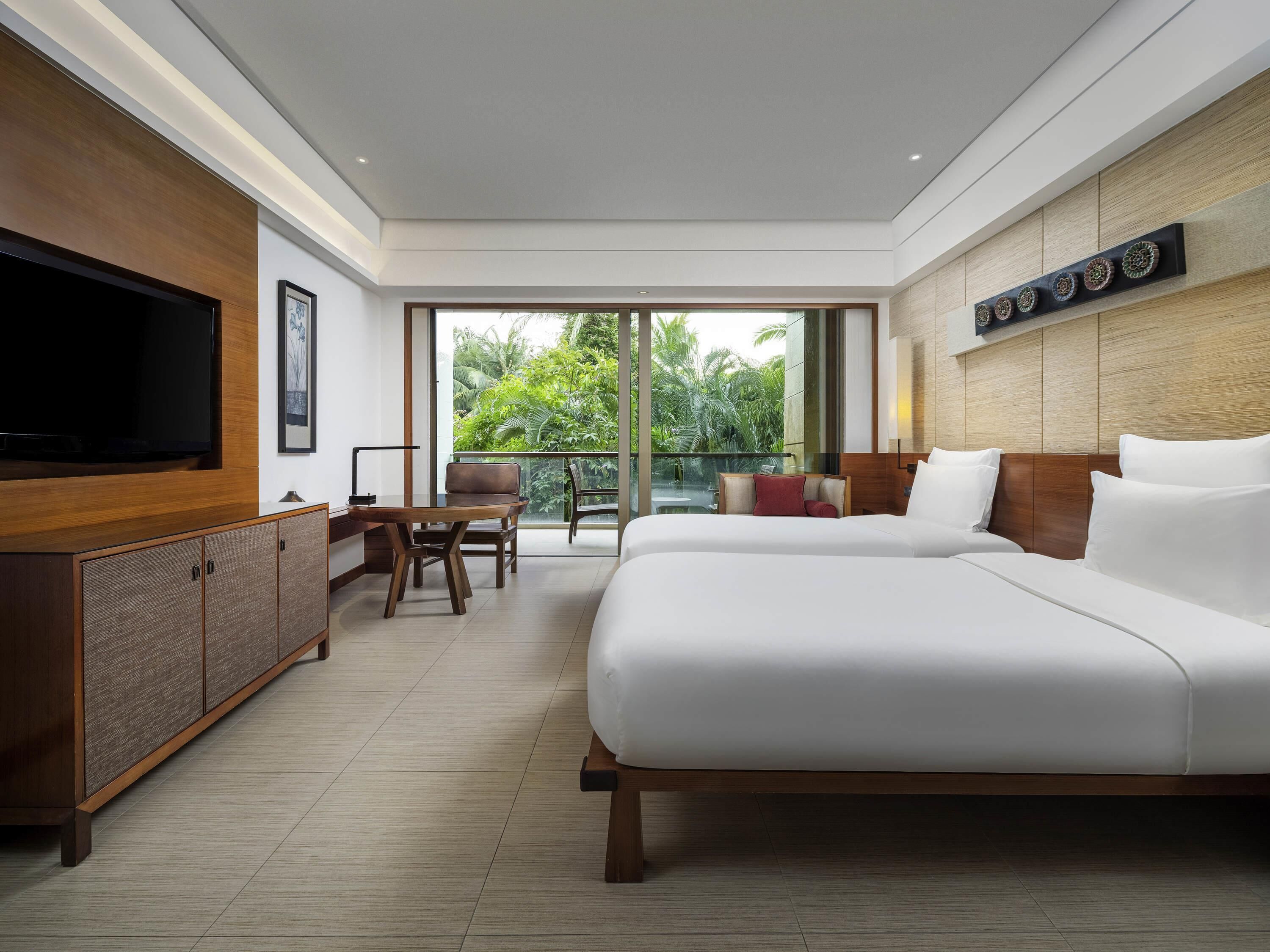 A modern hotel room with a large bed, a TV, a desk, and a balcony with a view of greenery.