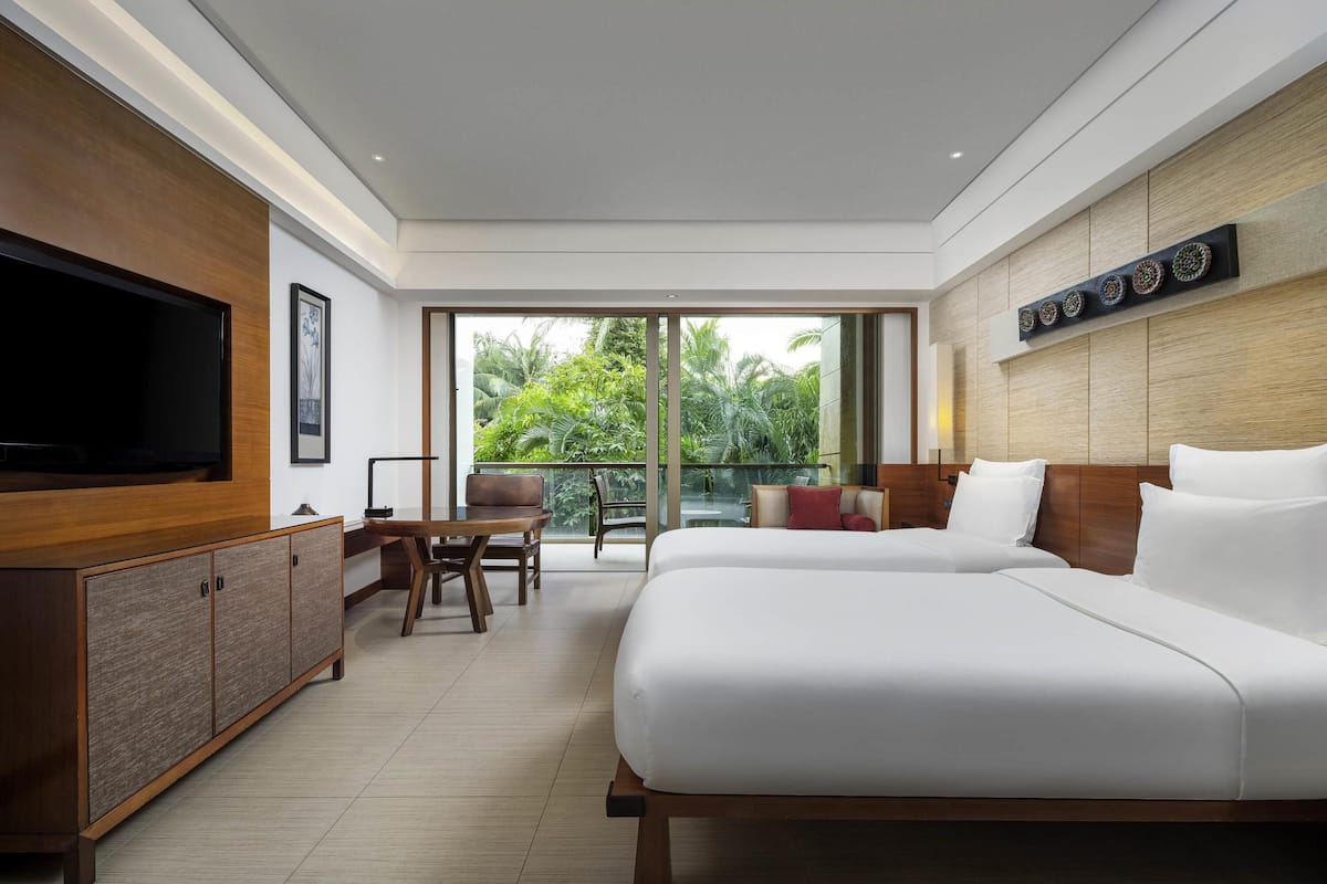 A modern hotel room with a large bed, a TV, a desk, and a balcony with a view of greenery.
