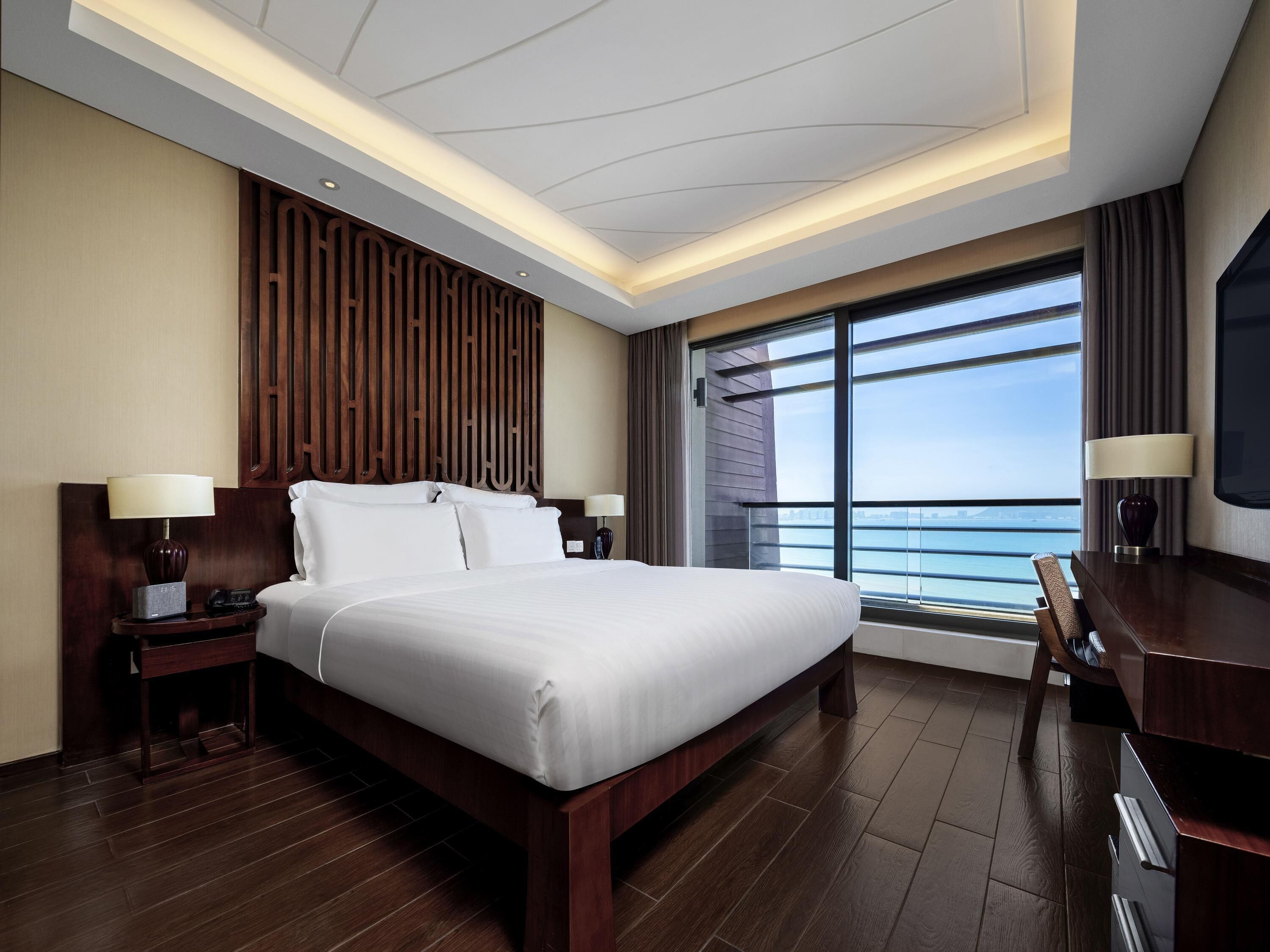 A hotel room with a large bed, wooden headboard, bedside tables with lamps, a TV, and a large window with a view of the sea.