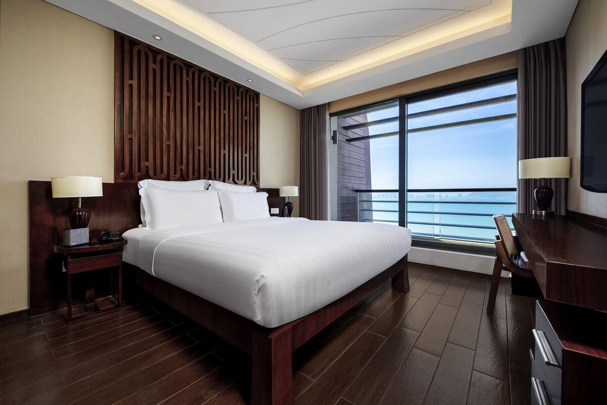 A hotel room with a large bed, wooden headboard, bedside tables with lamps, a TV, and a large window with a view of the sea.