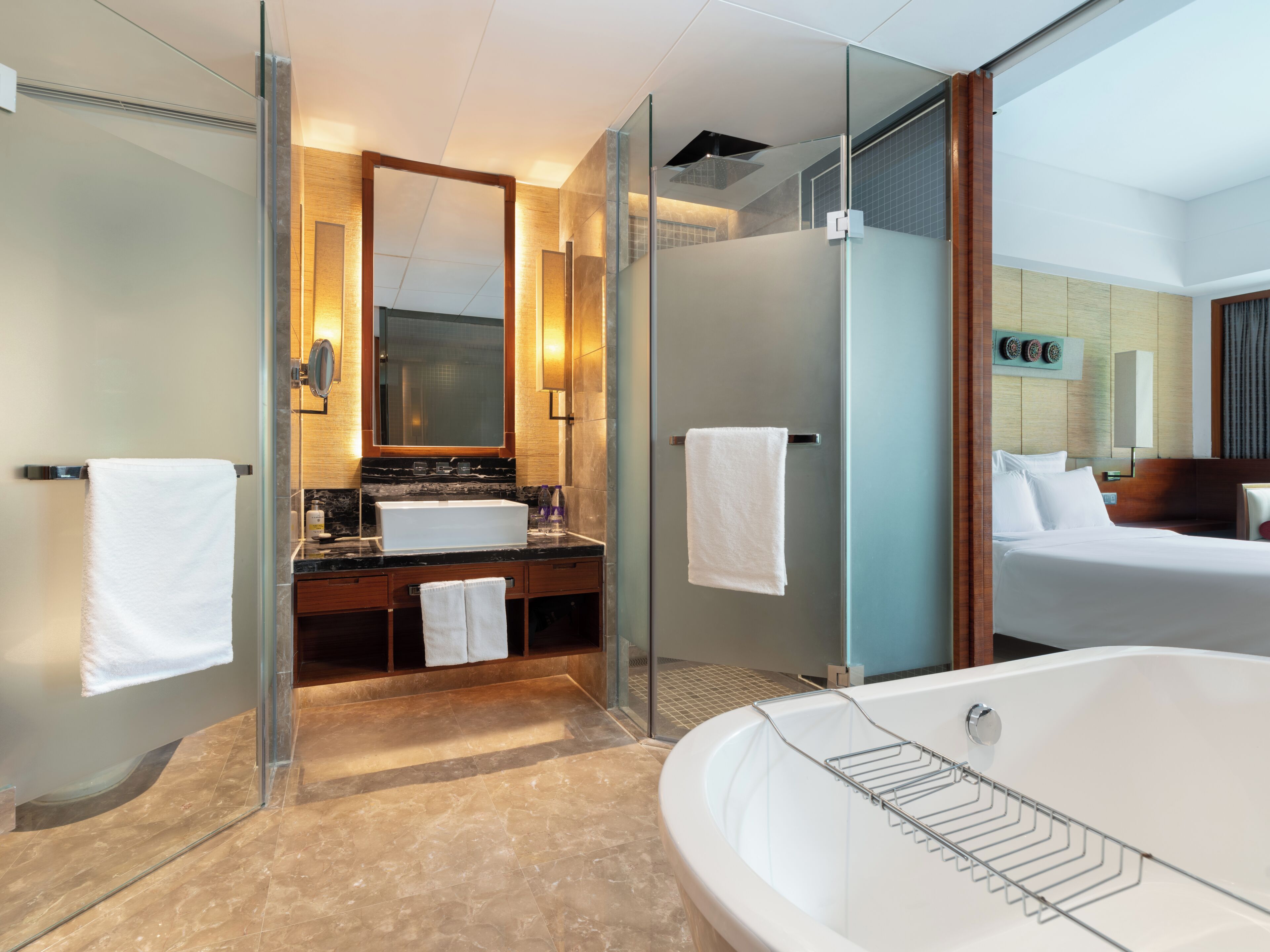 A modern hotel bathroom with a glass-enclosed shower, a large bathtub, a vanity with a mirror, and a bed in the adjacent room.
