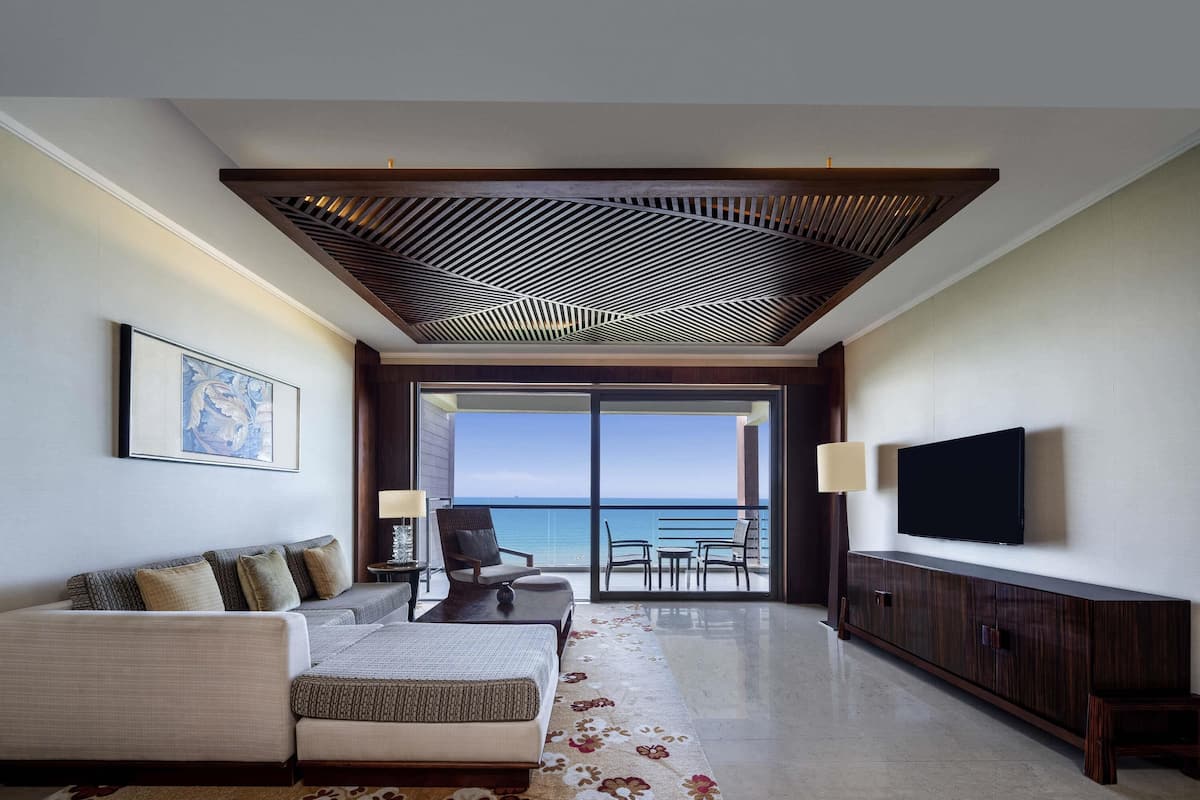 A modern hotel room with a balcony, a sofa, a TV, and a large window offering a view of the sea.