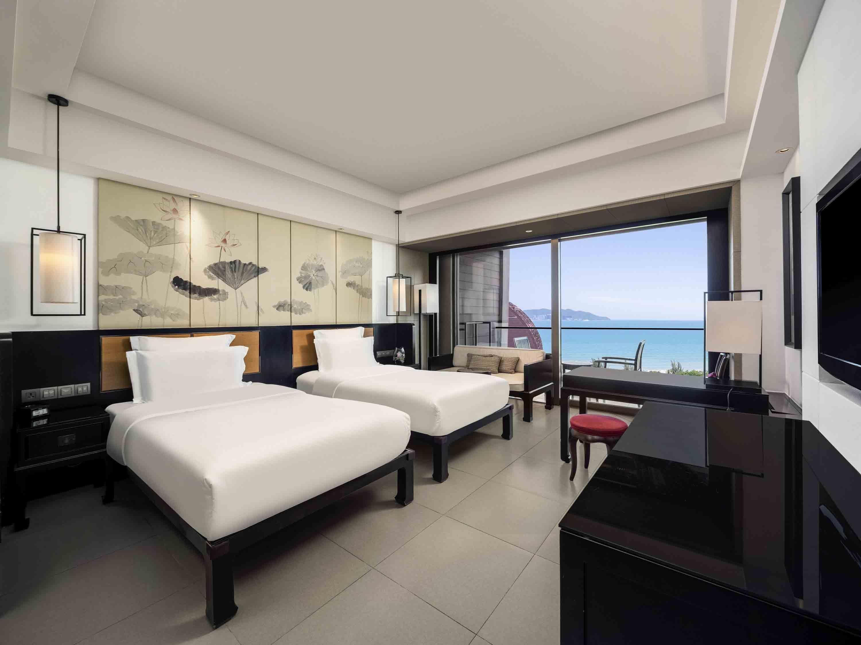 A modern hotel room with two beds, a desk, a TV, and a large window with a view of the sea.