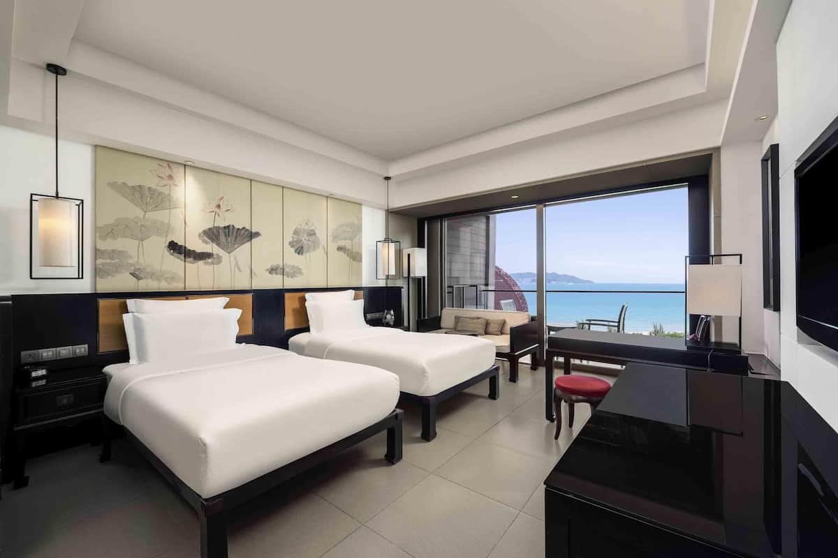 A modern hotel room with two beds, a desk, a TV, and a large window with a view of the sea.
