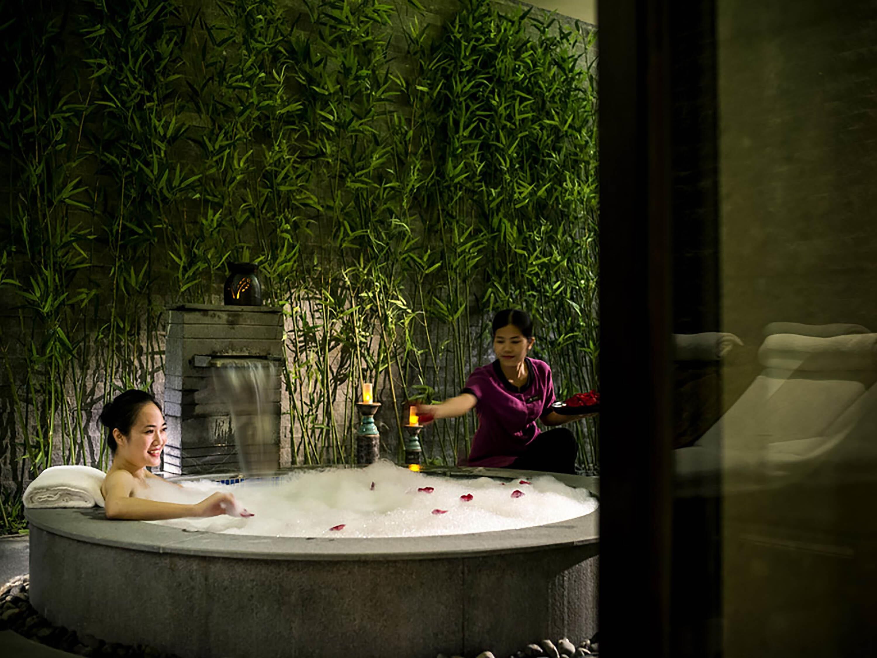 A spa setting with a hot tub, bamboo wall, and a person serving another individual.