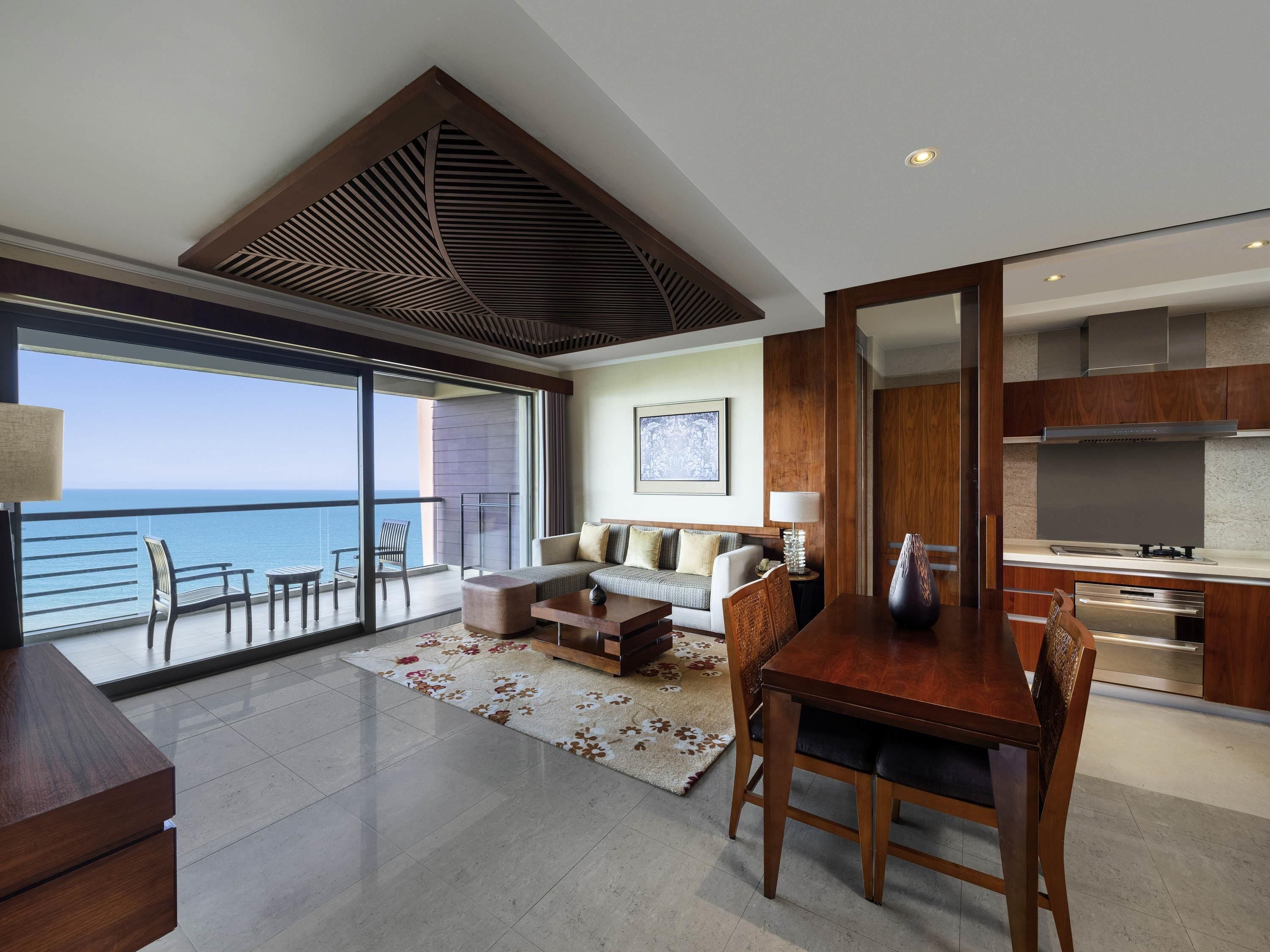 A modern living room with a balcony overlooking the sea, a dining area, and a kitchen with stainless steel appliances.