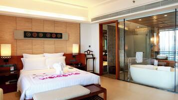 A hotel room with a large bed, bedside tables, a bench, a glass-enclosed bathroom with a bathtub, and a wooden headboard.