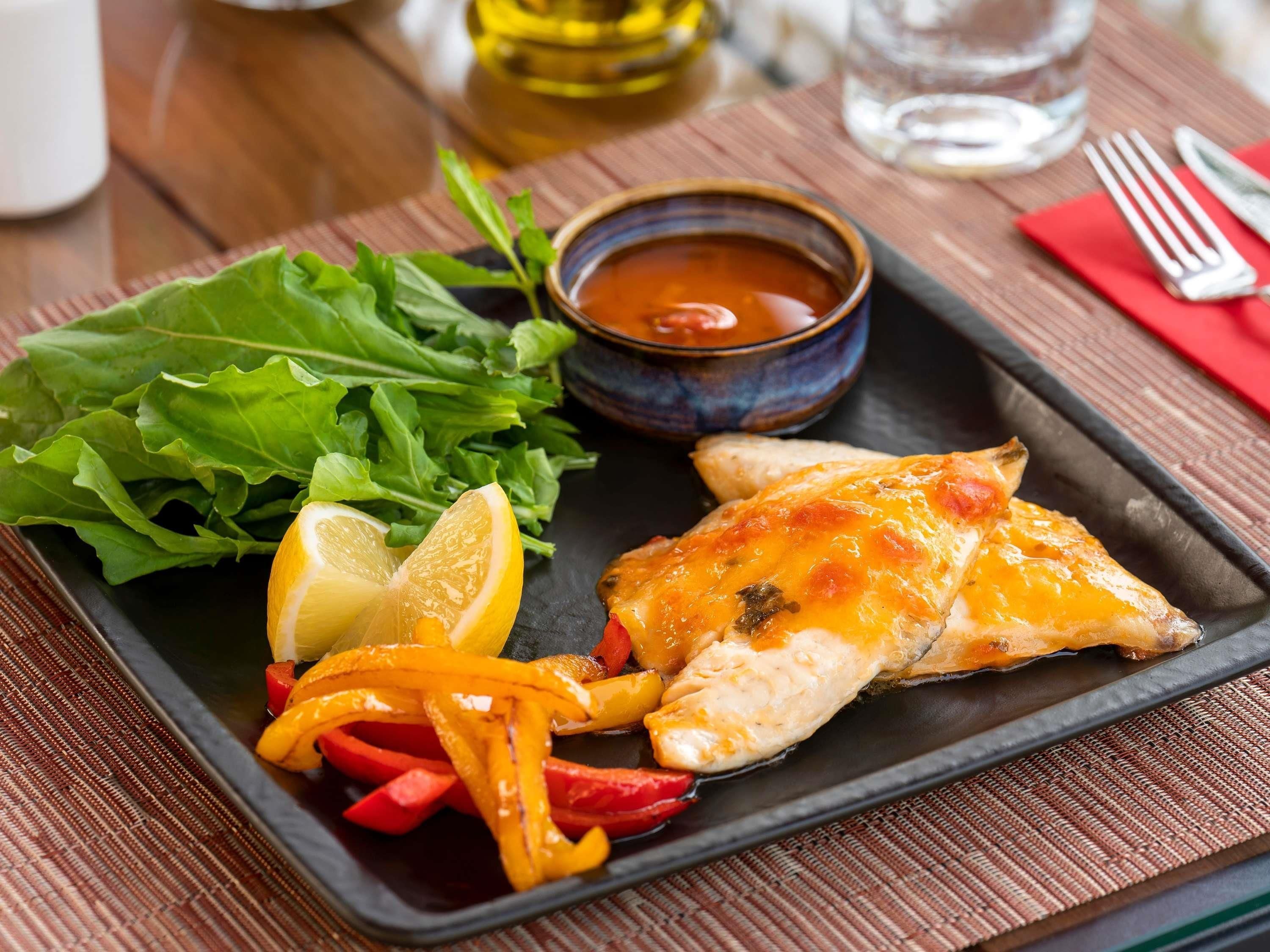 A meal with grilled chicken, lemon slices, bell peppers, and greens on a black plate, with a bowl of sauce and cutlery on a wooden table.