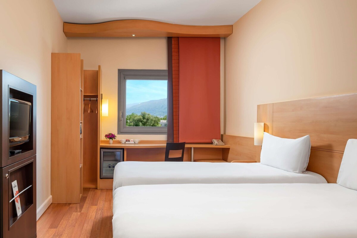 A hotel room with two beds, a desk with a chair, a TV, a window with a view, and a minibar.