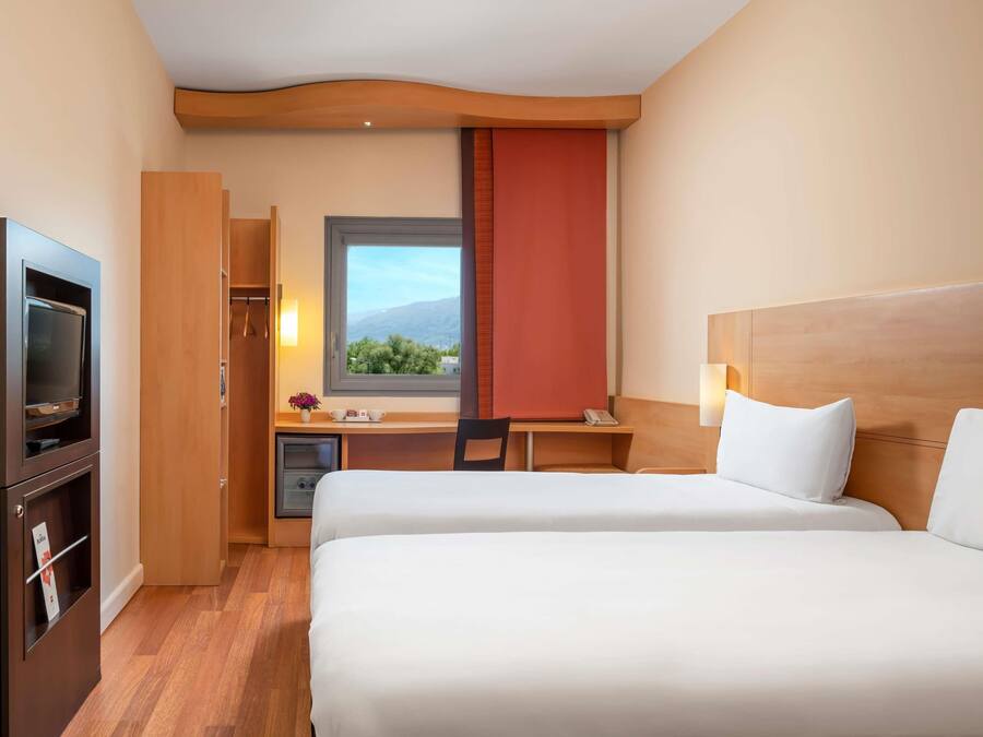 A hotel room with two beds, a desk with a chair, a TV, a window with a view, and a minibar.