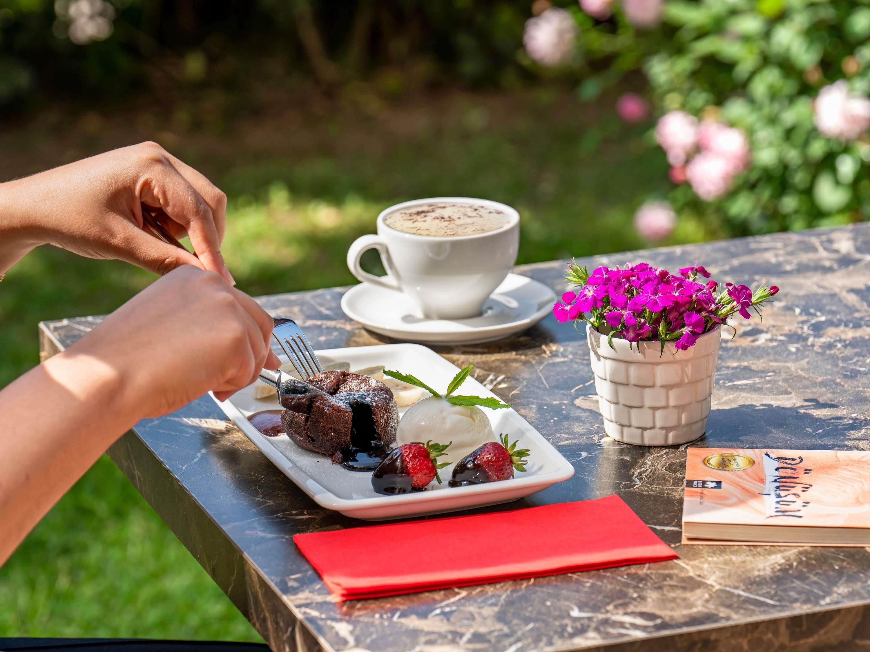 Eine Person isst draußen, mit einem Teller Essen, einer Kaffeetasse und einem Buch auf dem Tisch.