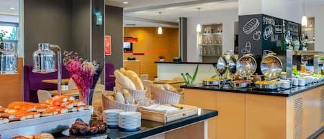 Zona de bufé con diferentes opciones de comida, área de comedor con sillas moradas y mostrador con menú en una pizarra.