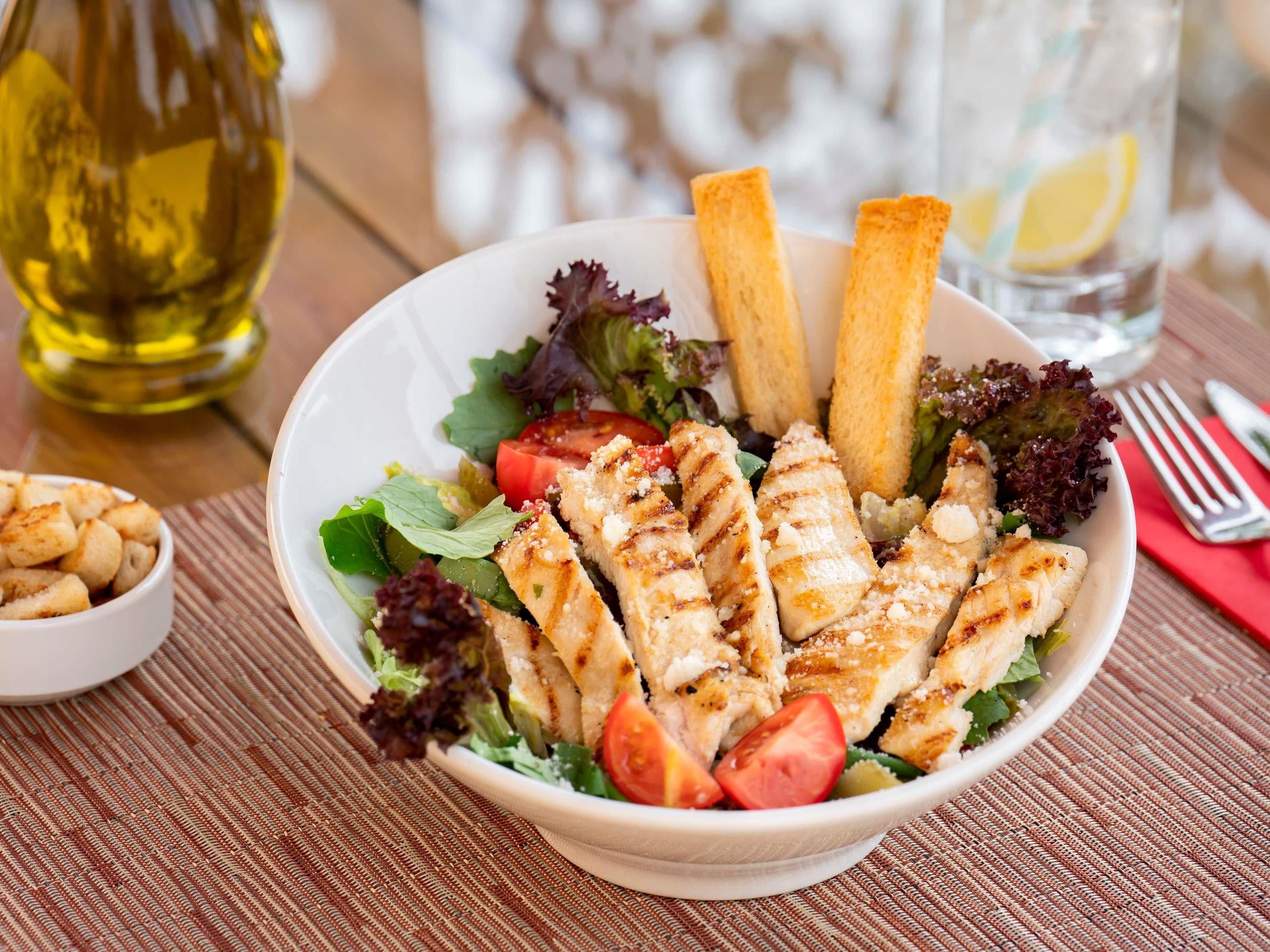 Un tazón de ensalada de pollo a la parrilla con lechuga, tomates y croutons, acompañado de una botella de aceite de oliva y un vaso de agua.