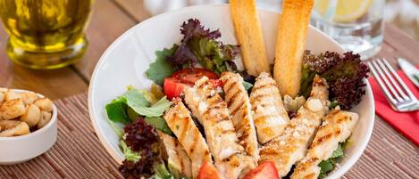 Un tazón de ensalada de pollo a la parrilla con lechuga, tomates y croutons, acompañado de una botella de aceite de oliva y un vaso de agua.