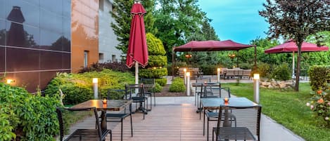An outdoor patio with tables and chairs, red umbrellas, and a building in the background.