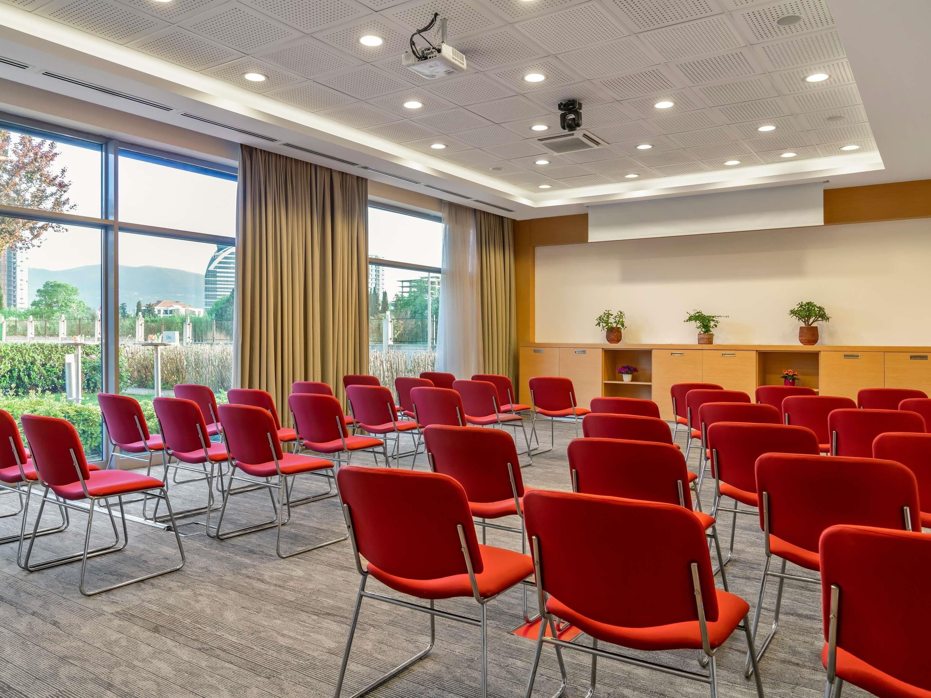 Un salón de conferencias con sillas rojas, un proyector y vistas al exterior con vegetación.