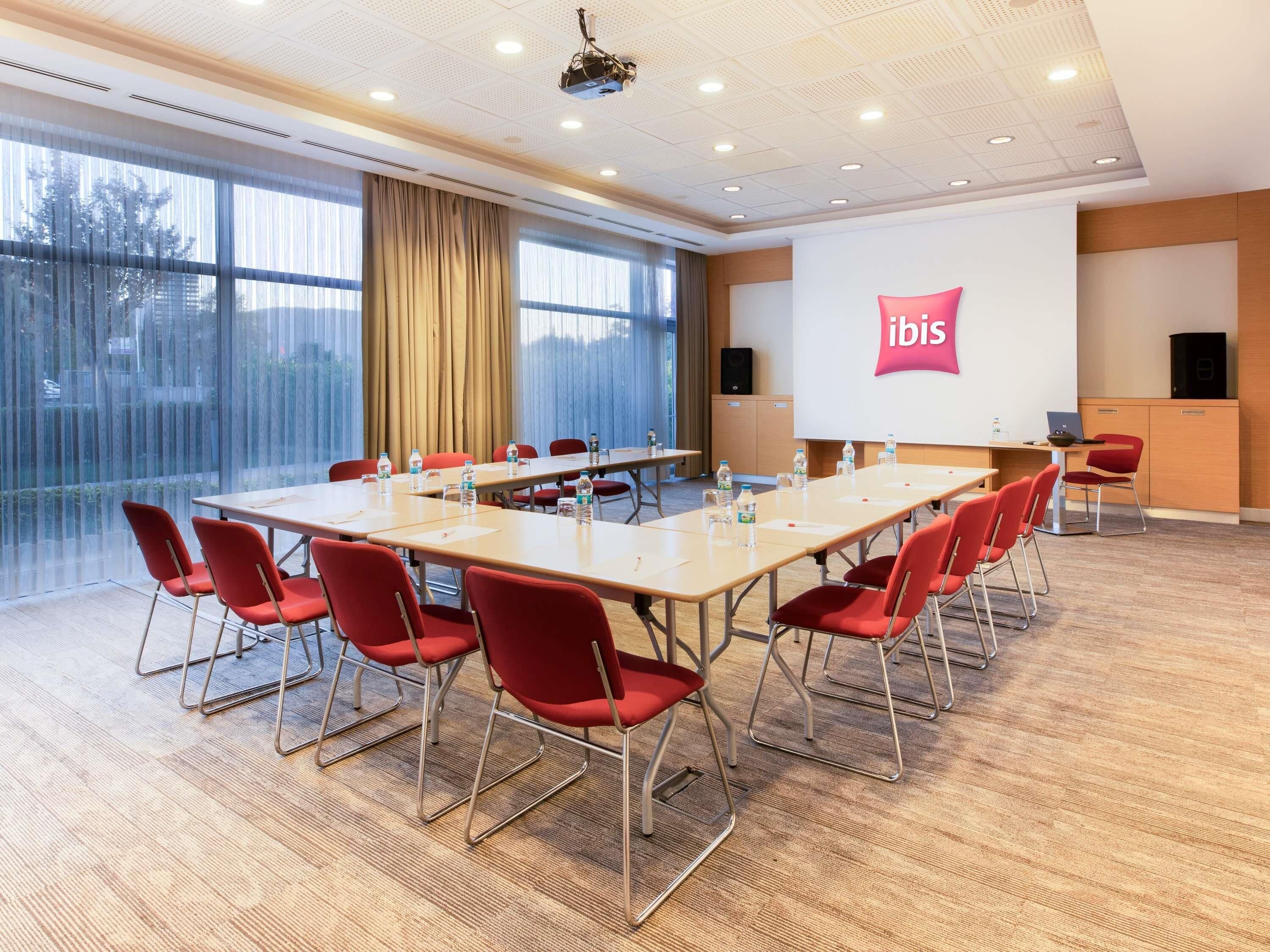 A conference room with a long table, red chairs, a projector screen, and a large window with curtains.