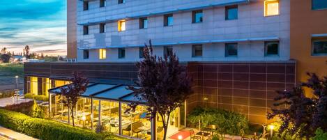 A multi-story hotel with a glass facade, outdoor seating area, and a parking lot.