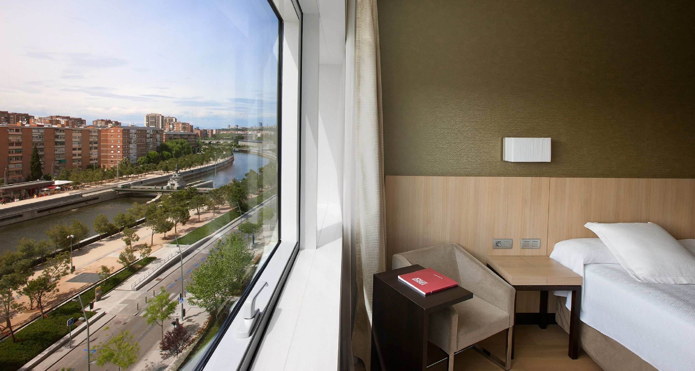 A hotel room with a large window overlooking a river and cityscape, a bed, a small table with a book, and a wall-mounted light.