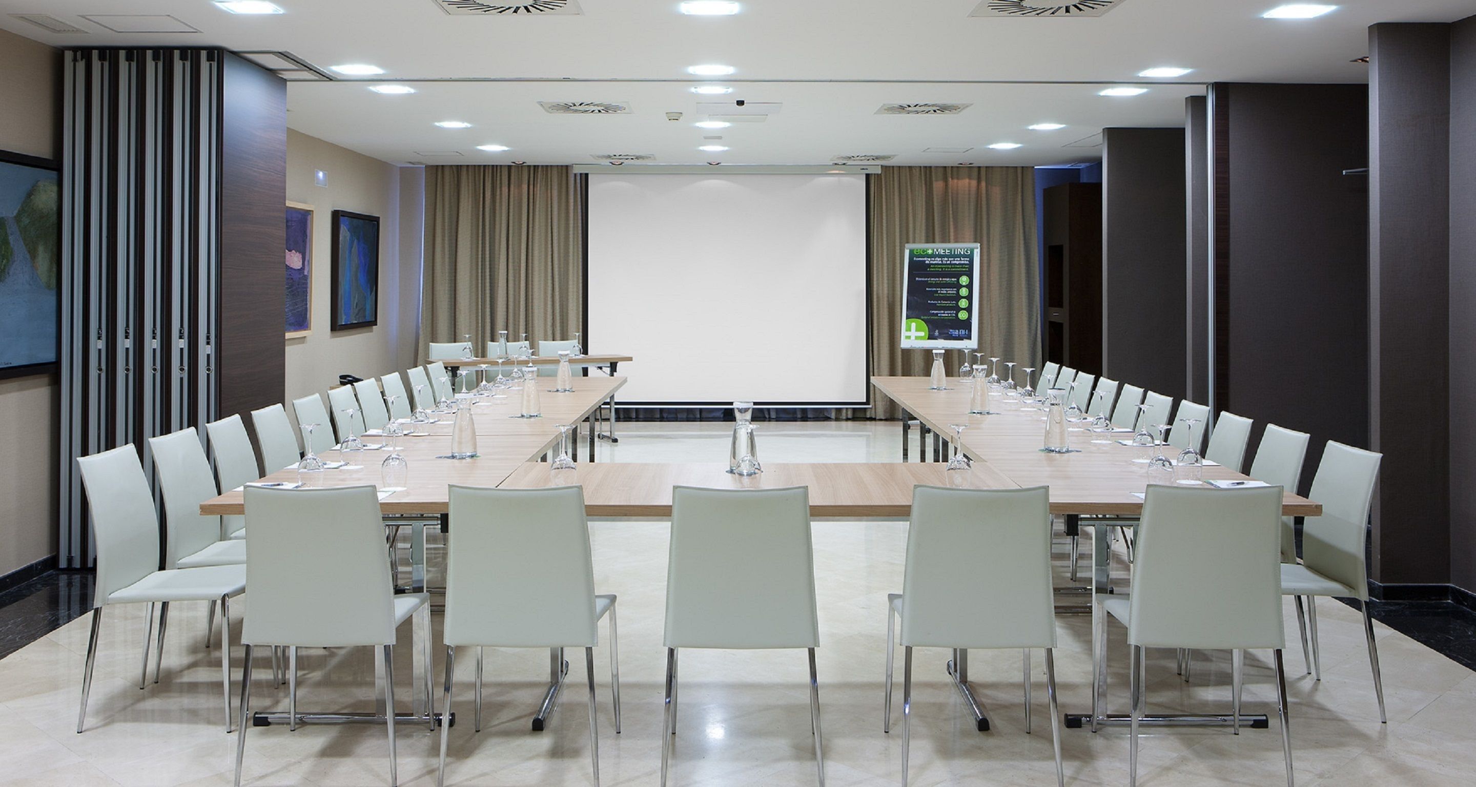 Un salón de conferencias con una larga mesa, sillas blancas y una pantalla de proyector.
