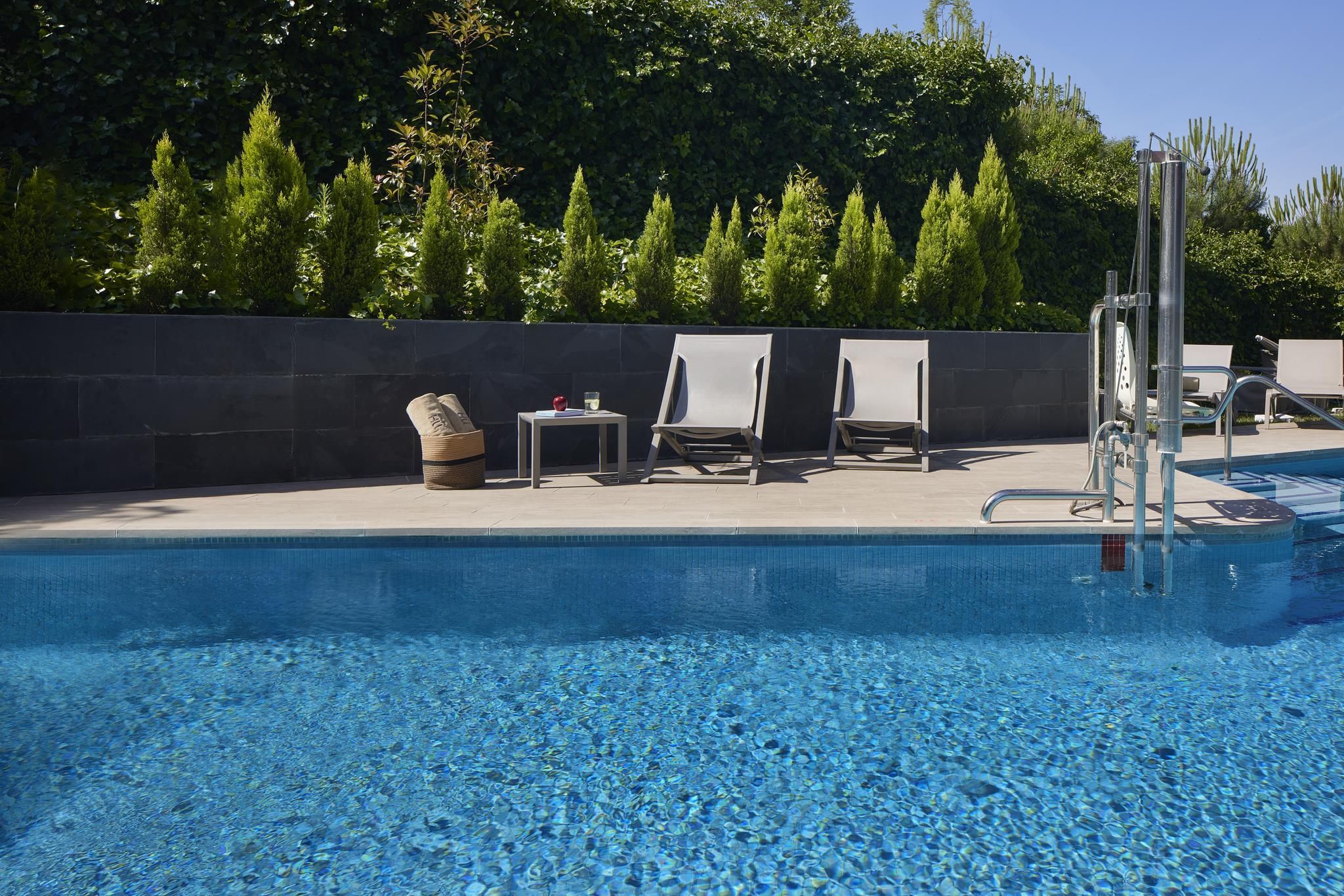 Una piscina con agua cristalina azul, rodeada de sillas de descanso y un muro de piedra.