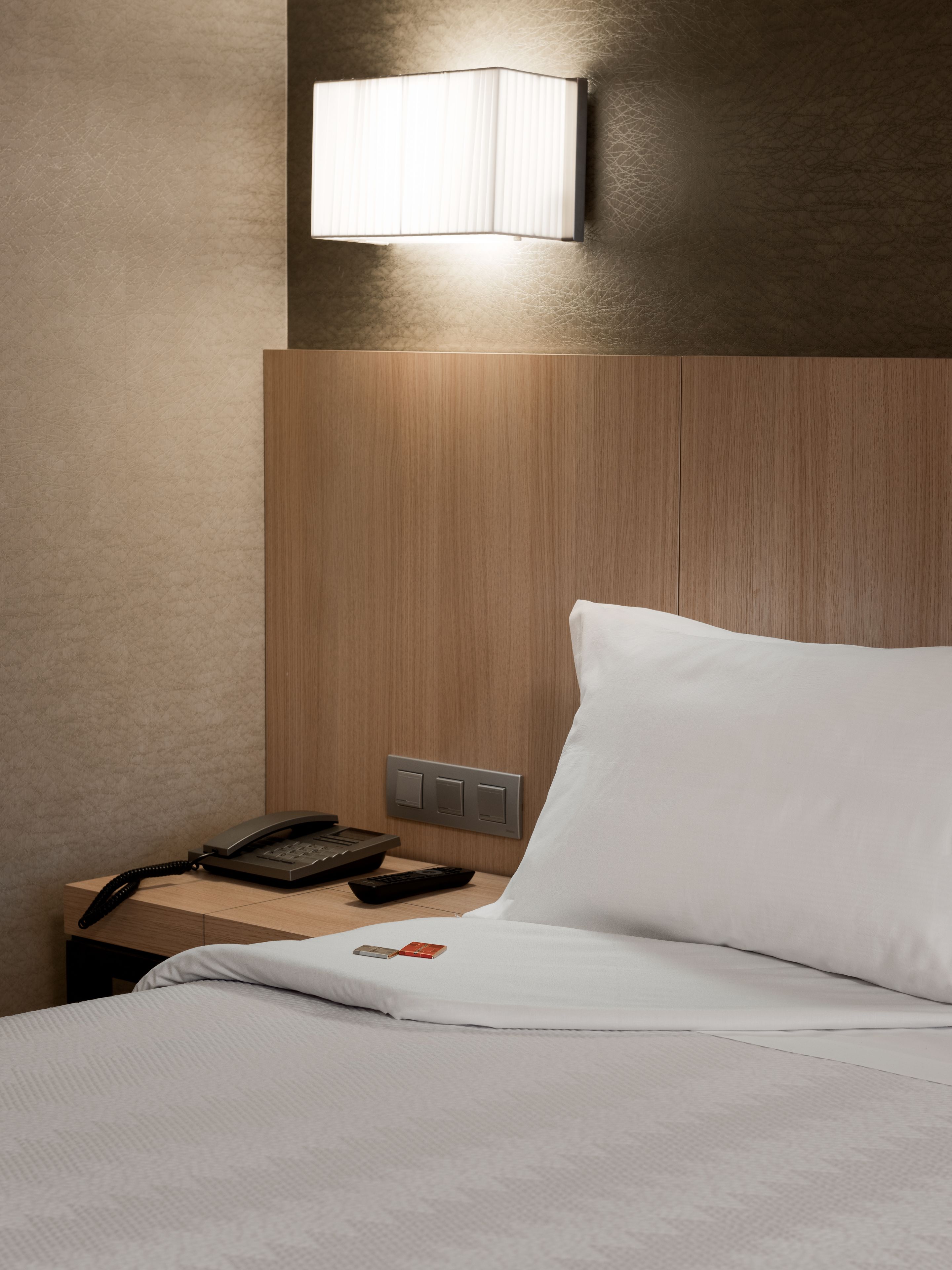 Habitación de hotel con cabecera de madera, mesita de noche con teléfono y control remoto, almohada blanca y lámpara de pared.