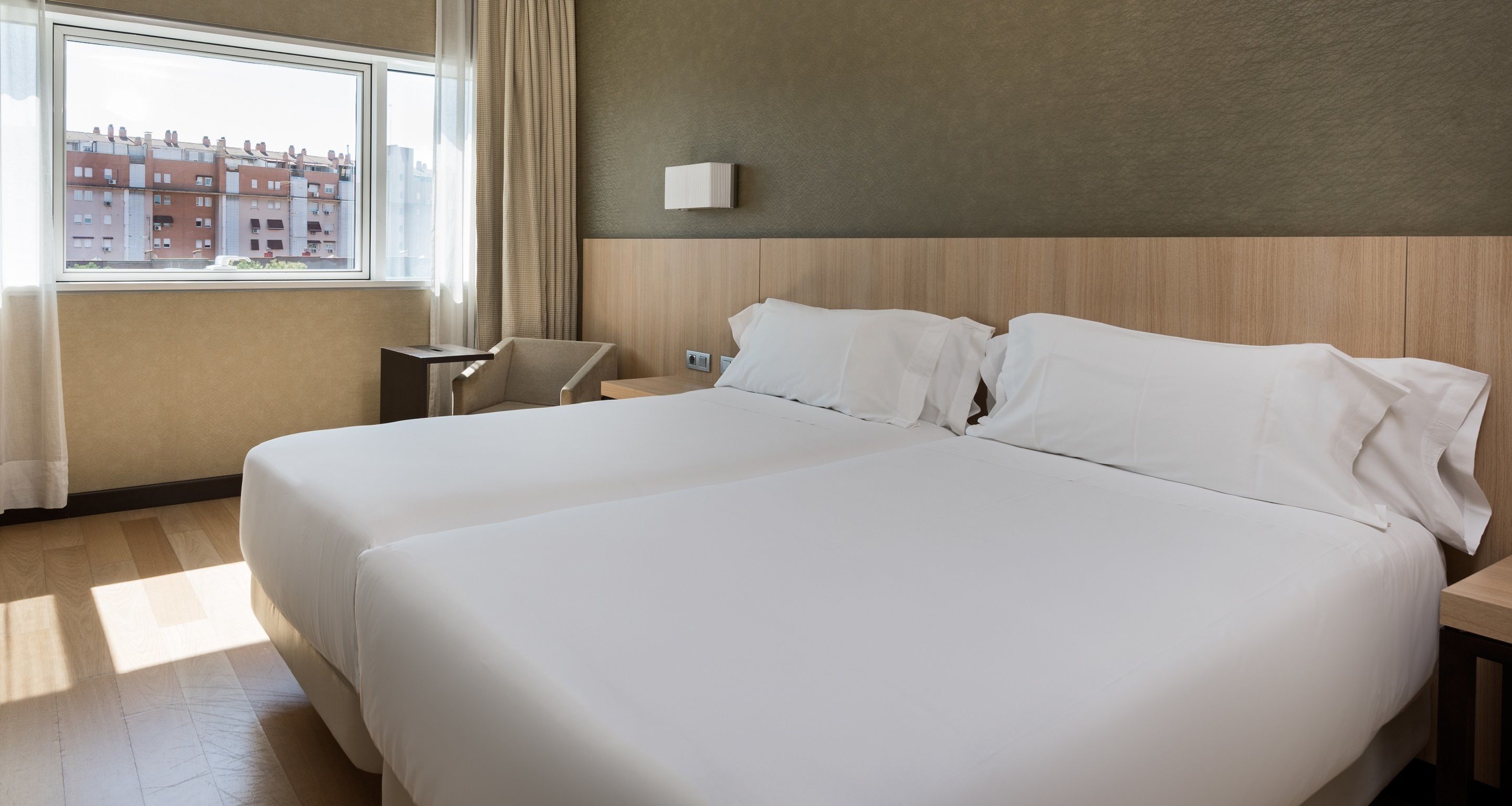 A hotel room with two large beds, a small table, a chair, and a window with curtains.