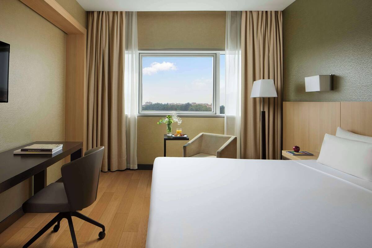 Una habitación de hotel con cama, escritorio, silla, lámpara y ventana con cortinas.