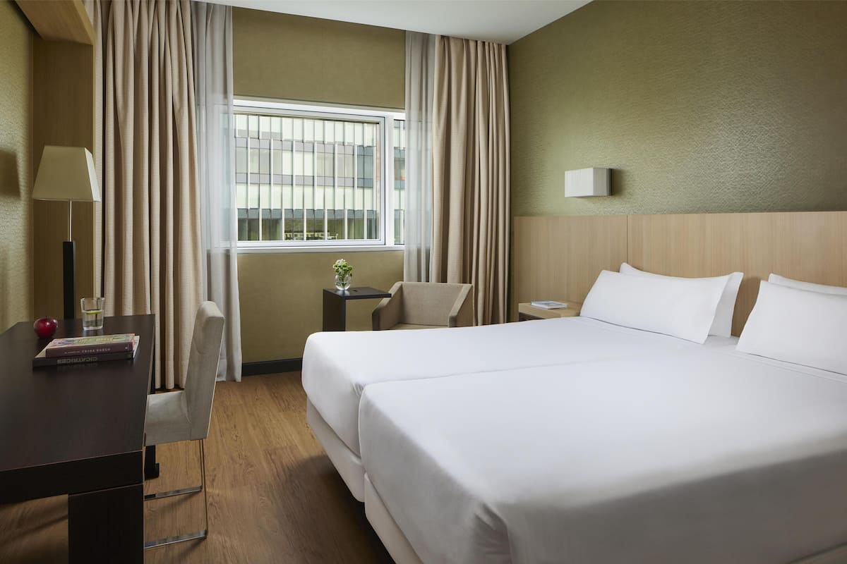 Habitación de hotel con una cama grande, un escritorio con lámpara, una silla, una mesita con un jarrón y una ventana con cortinas.