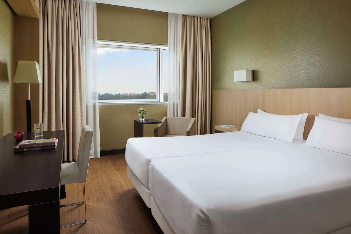 Una habitación de hotel con una cama grande, un escritorio con una lámpara, una silla y una mesita con un florero y un libro.