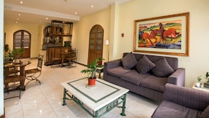 A spacious living area with a sofa, a dining table, and chairs. There is a kitchen in the background with wooden cabinets and a large painting on the wall. - RQ Hotel Dacarlo (Santiago)