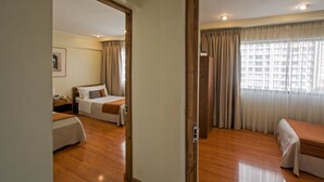 A hotel room with a wooden floor, a bed with a white comforter, a bedside table with a lamp, and a window with curtains. - RQ Hotel Dacarlo (Santiago)