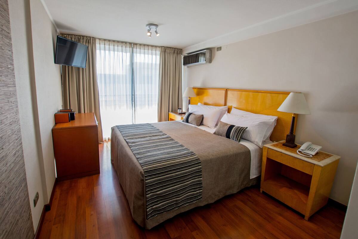 Ein Hotelzimmer mit einem Bett, einem an der Wand befestigten Fernseher, einem Nachttisch mit Lampe und Telefon sowie einem Fenster mit Vorhängen.