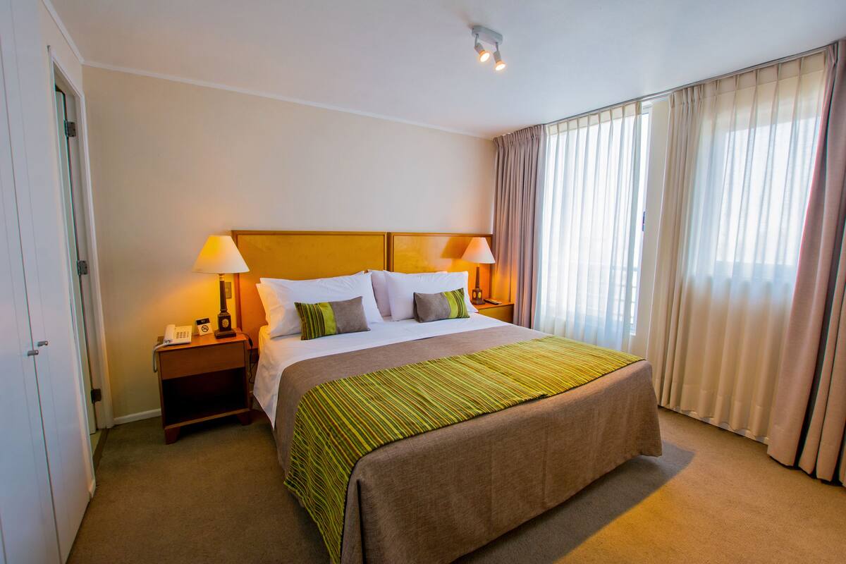 Ein Hotelzimmer mit einem großen Bett, zwei Nachttischlampen, einem Telefon und einem fenster mit Vorhängen.