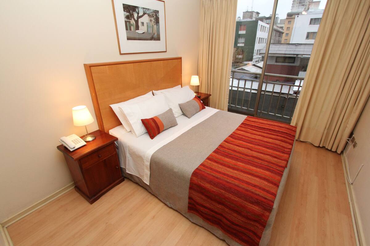 A hotel room with a bed, bedside lamps, a telephone, a window with curtains, and a framed picture on the wall.