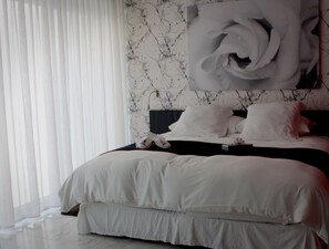 A bedroom with a large bed, white bedding, a headboard with a rose design, and a window with sheer curtains.