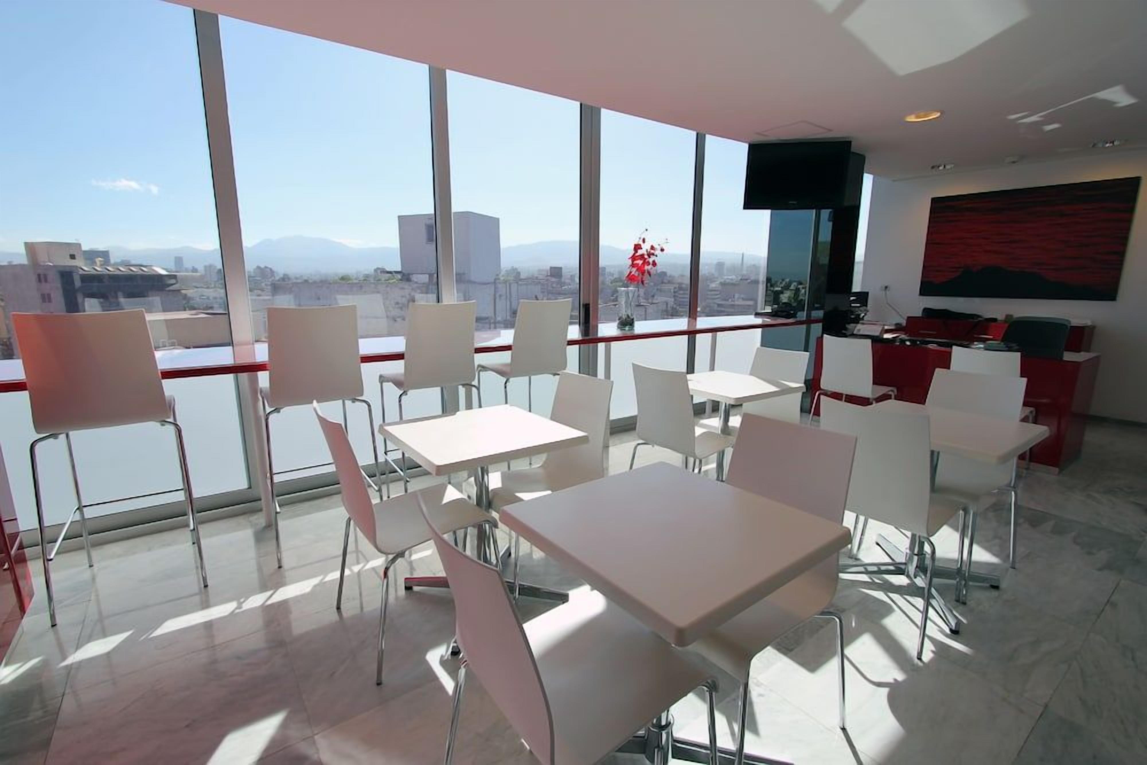 Un moderno comedor con mesas y sillas, ventanas amplias con vista a la ciudad y un televisor de pantalla plana montado en la pared.