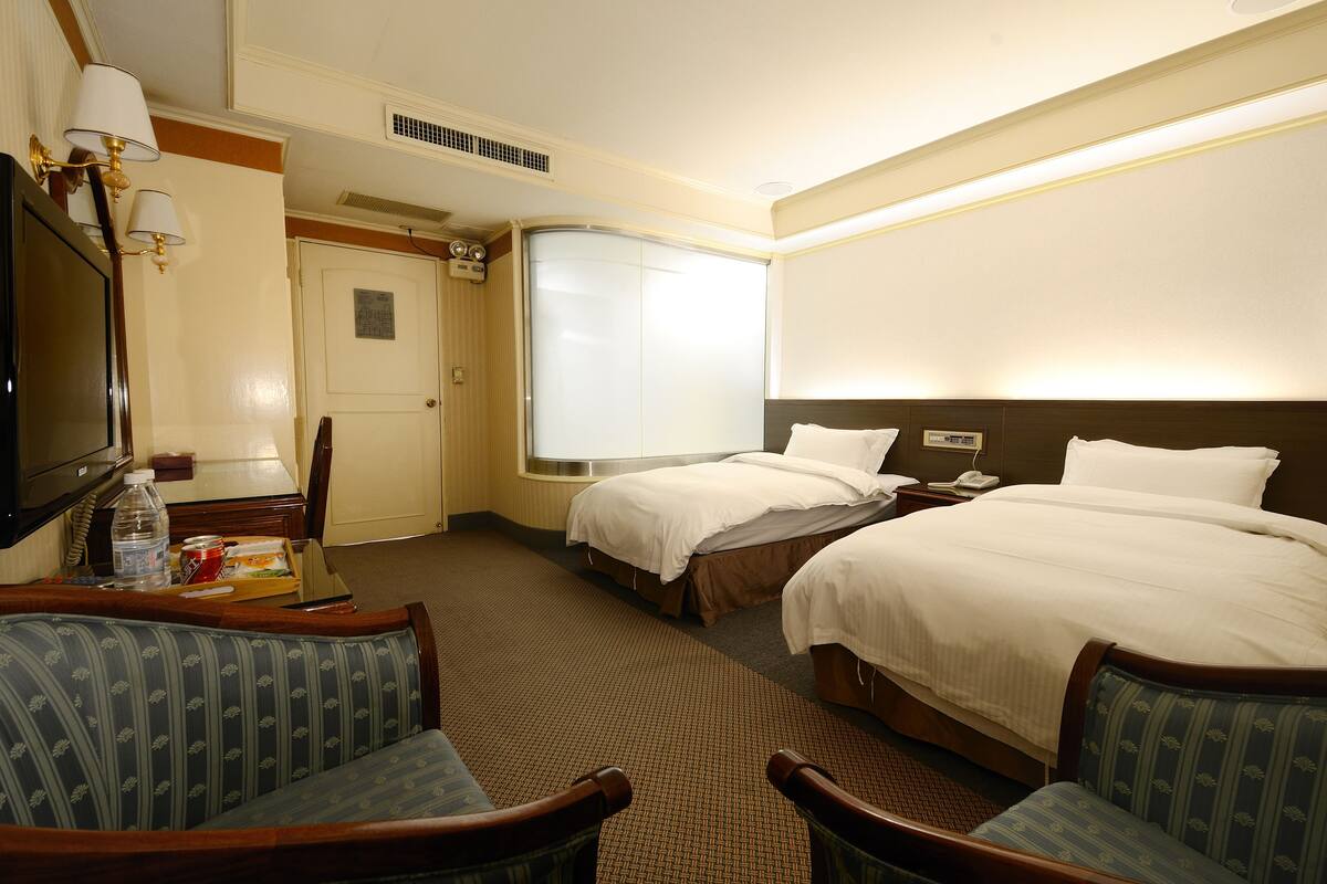 Une chambre d’hôtel avec deux lits, un bureau avec une chaise, une télévision et une fenêtre avec des stores.