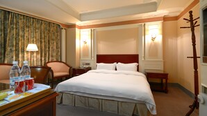 A hotel room with a large bed, two bedside tables, a desk with a chair, a lamp, and a coat rack.