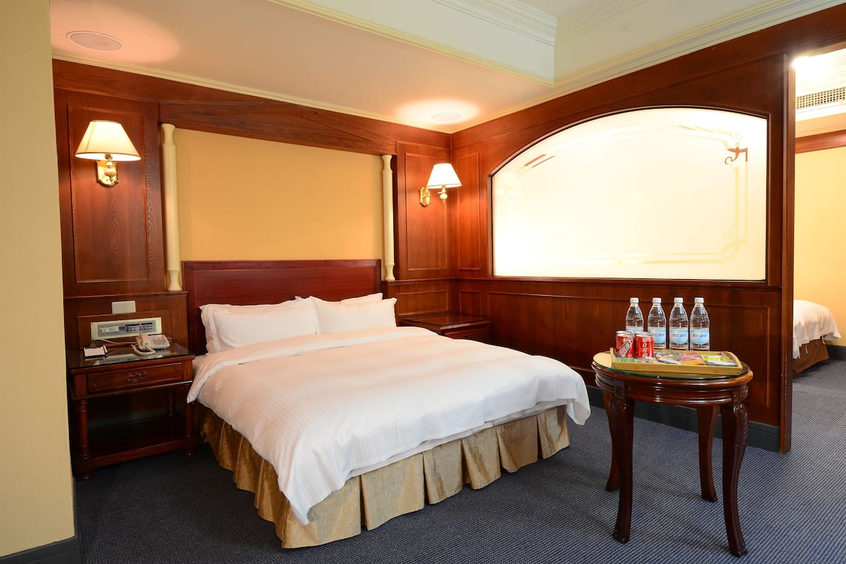 Une chambre d’hôtel avec un grand lit, une tête de lit en bois, deux lampes de chevet, une table de chevet avec un téléphone et une petite table avec des bouteilles d’eau et des en-cas.