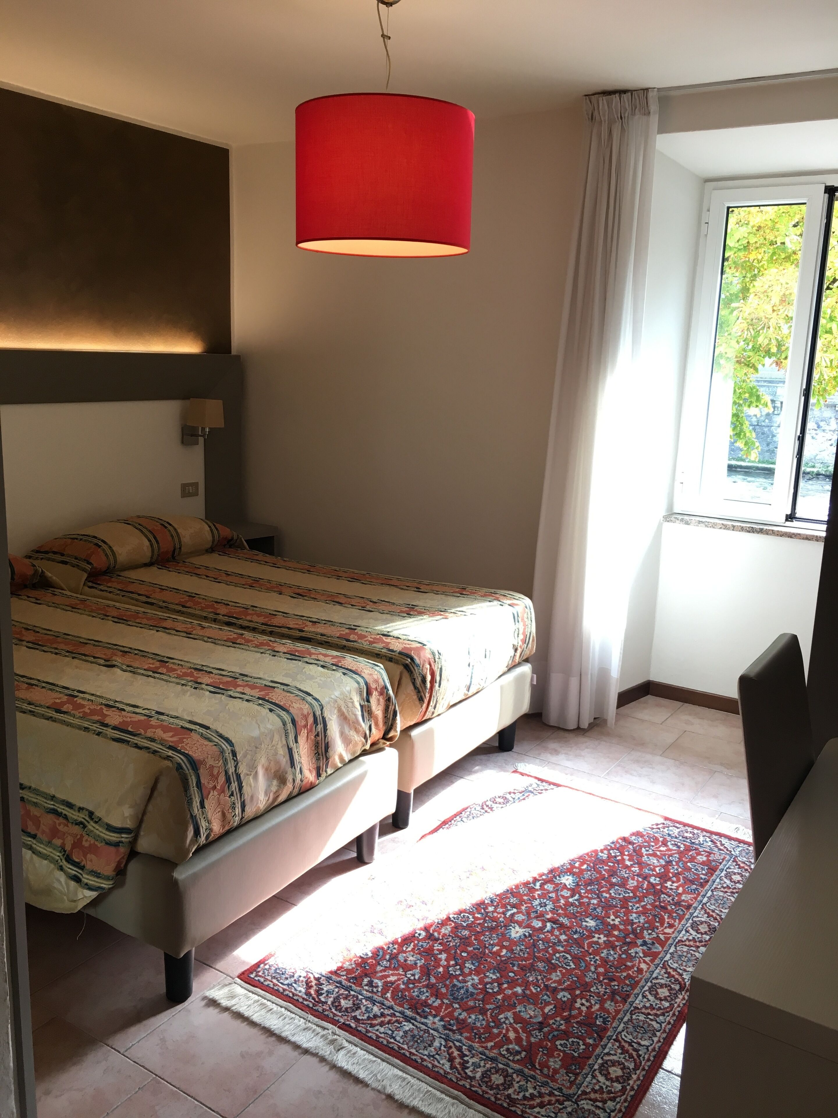 A bedroom with a bed, a red hanging lamp, a window with curtains, and a patterned rug.