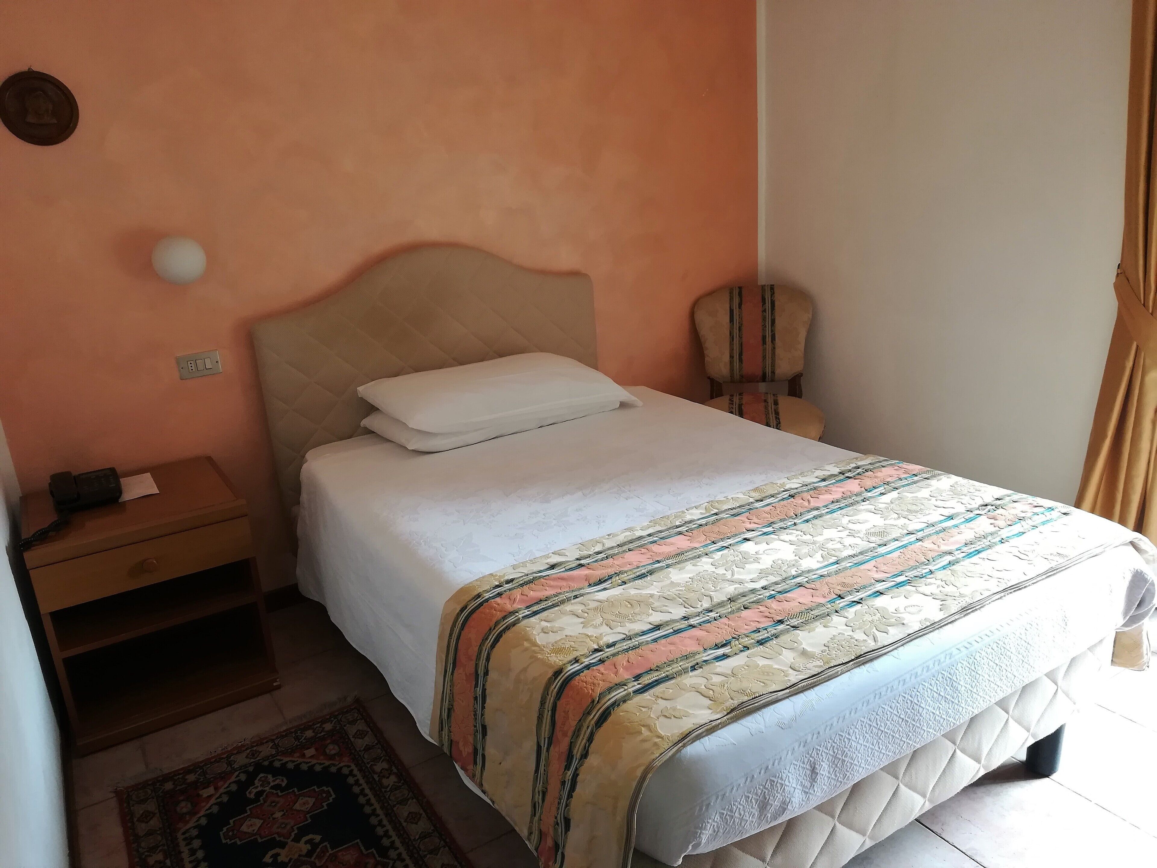 A neatly made bed with a patterned bedspread, a bedside table with a lamp, and a chair against the wall.