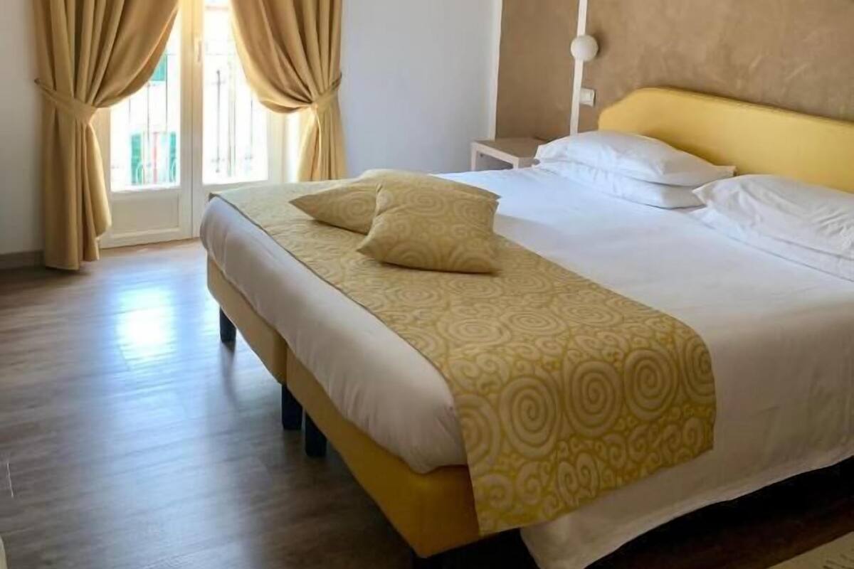 Ein Hotelzimmer mit einem großen Bett, beigen Vorhängen, einer weißen Deckenleuchte und einem Holzboden.