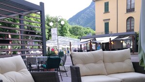 Un espace extérieur aménagé avec des meubles en osier et une table en verre, entouré de bâtiments et avec une montagne en arrière-plan.